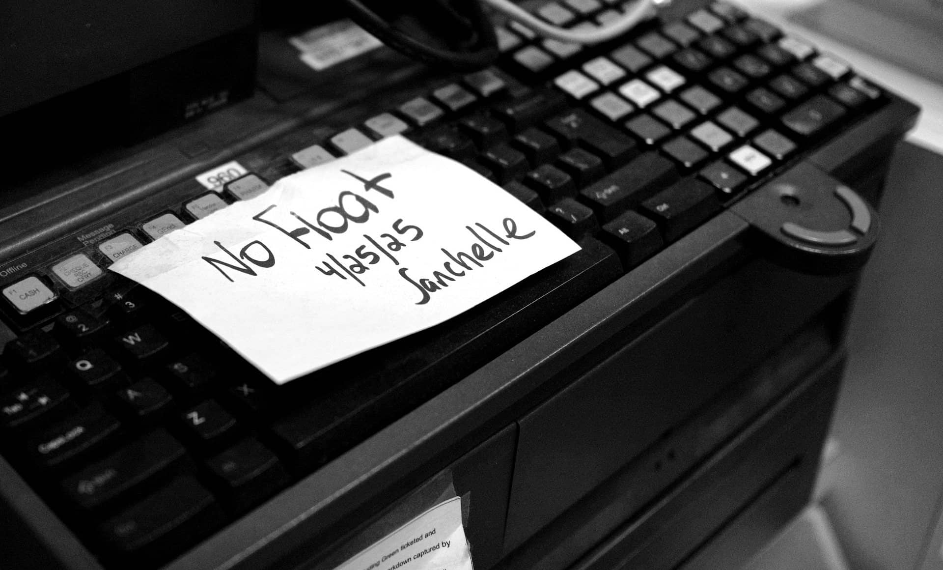 A shop till, photographed close: worn keys and a handwritten no-float note taped over them.