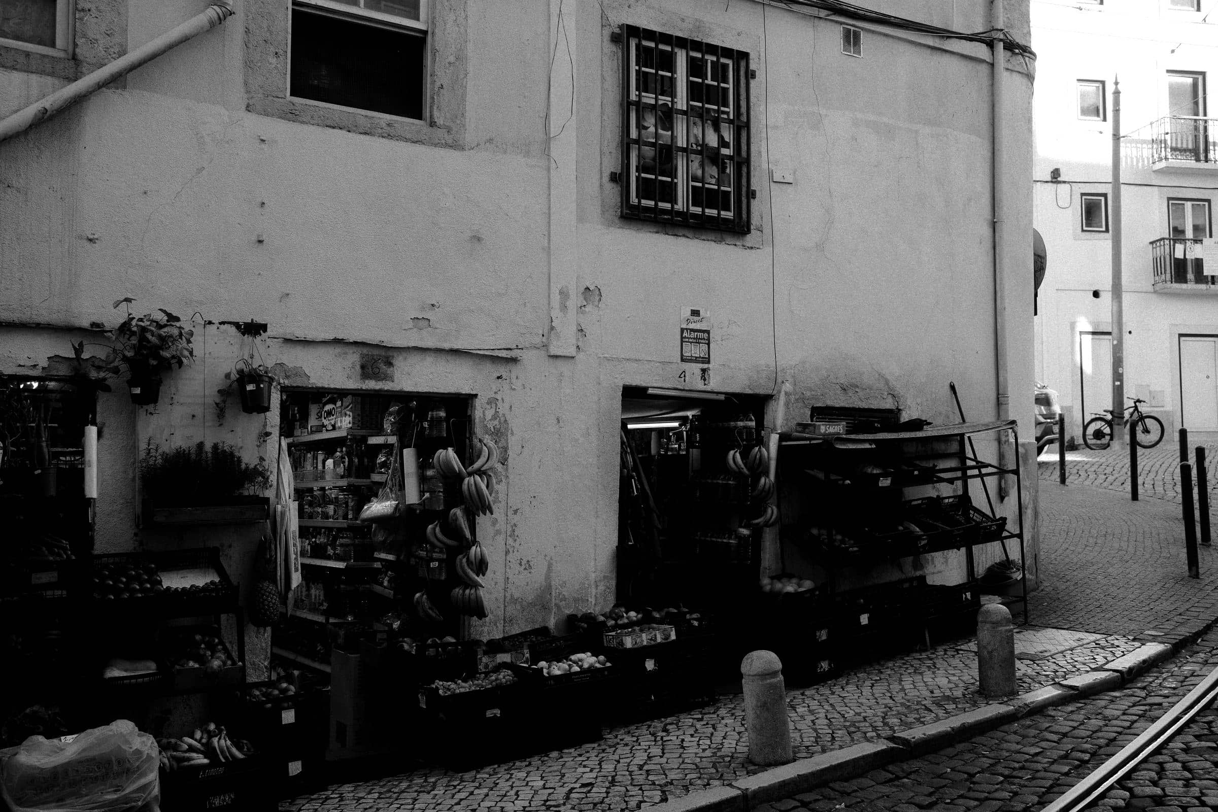 Um minimercado de esquina em Lisboa: caixas de fruta empilhadas no passeio, calçada portuguesa, ninguém à porta.