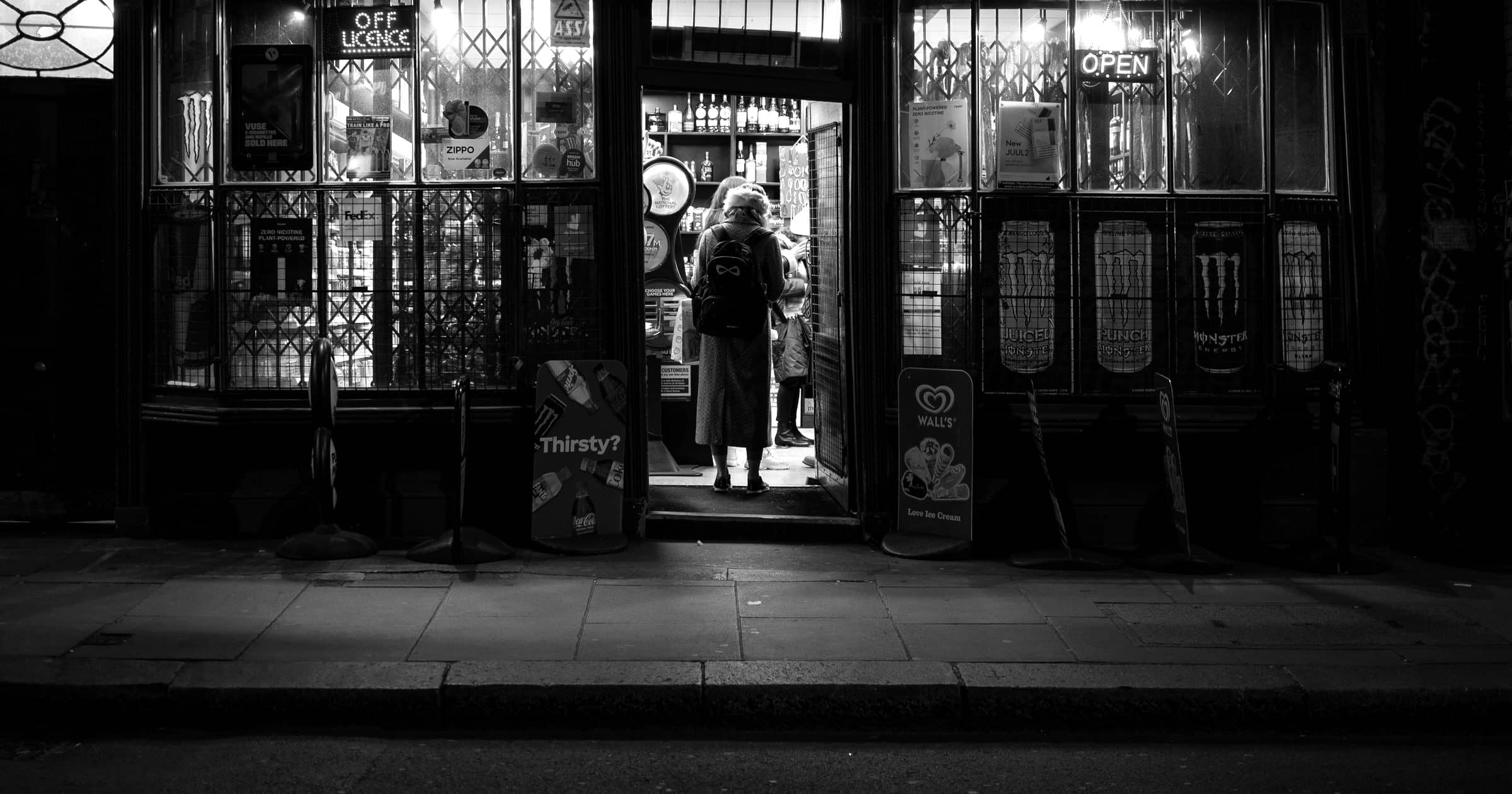 The lit doorway of a newsagent and off licence at night, one person in the doorway seen from behind.